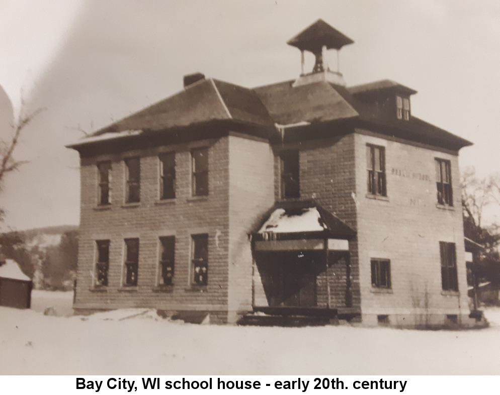 Black and white photo of a two-story brick school house with hip roofs and a belfry. It is winter and snow is on the ground and on the roof of the small entry porch under a grey sky.